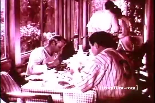The still features a vintage dining scene, likely from a 1970s Kellogg's Corn Flakes advertisement. It shows two men seated at a table with a checkered tablecloth, enjoying breakfast. The setting appears to be a sunlit room with large windows, suggesting a casual, relaxed atmosphere. The focus is on their meal, emphasizing the product. One man wears a baseball jersey, hinting at a connection to the sport, possibly Babe Ruth. The overall tone is nostalgic and embraces a classic Americana feel.