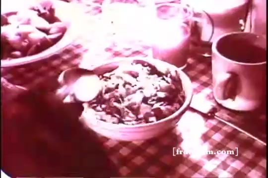 The still features a close-up of a bowl of Kellogg's Corn Flakes, with a spoon in motion, set against a checkered tablecloth. Surrounding the bowl are a glass of milk and a mug, with a background hinting at an abundance of food items, creating a wholesome breakfast scene. The vintage 16mm print gives it a warm, nostalgic feel.