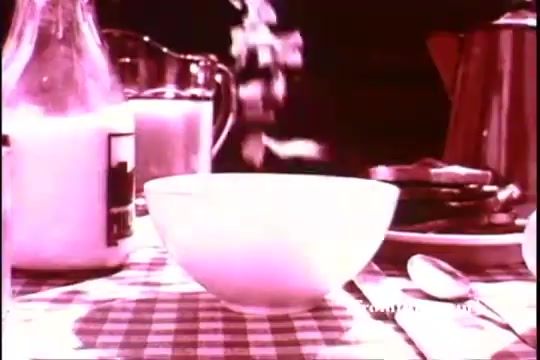 The still from the 1970s Kellogg's Corn Flakes commercial features a breakfast scene with a focus on a white bowl as corn flakes are poured into it. There is milk in a glass bottle nearby, and other breakfast items, like pancakes, can be seen on a checkered tablecloth. The overall mood suggests a wholesome morning routine, emphasizing the product as part of a classic American breakfast.