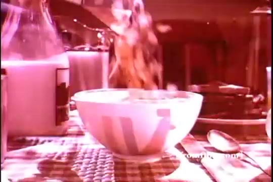 The still captures a vibrant scene from a 1970s Kellogg's Corn Flakes commercial, featuring a bowl being filled with corn flakes, likely in an inviting kitchen setting. The milk jug suggests a wholesome breakfast moment. The warm color palette evokes nostalgia, and the overall composition emphasizes the product's appeal.