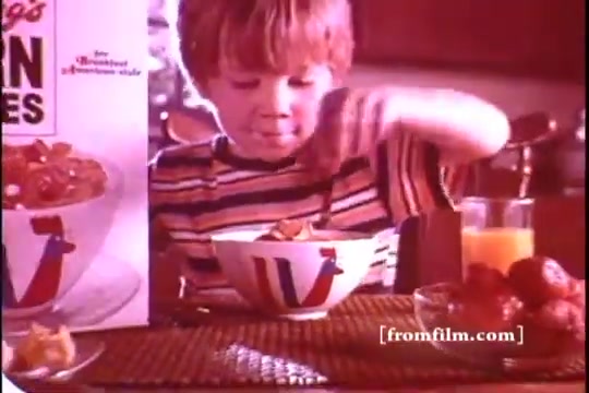 The still from the 1970s Kellogg's Corn Flakes commercial features a young boy with blonde hair wearing a striped shirt. He is intently eating from a bowl of cereal, with a box of Kellogg's Corn Flakes prominently displayed nearby. The setting is warm and homely, accompanied by fruits and a glass of orange juice, evoking a nostalgic breakfast scene. The color palette is rich, capturing the essence of classic commercials from that era.