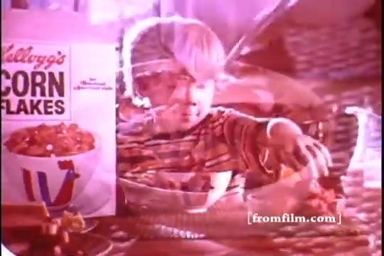 The still features a young child joyfully reaching into a bowl of Kellogg's Corn Flakes, with a prominent box of the cereal nearby. The colors are vibrant, typical of 1970s advertising. The nostalgic vibe captures the simplicity and joy associated with breakfast cereals in that era. The overall composition evokes a sense of warmth and childhood delight.