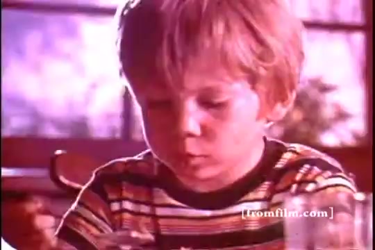 The still features a young boy with short, light-colored hair, looking intently at his bowl of cereal. He is wearing a striped shirt, indicative of the 1970s style. The warm lighting creates a nostalgic atmosphere. In the background, there are hints of a domestic kitchen setting, suggesting a familial breakfast scene. The context ties into a Kellogg's Corn Flakes advertisement, likely aiming to evoke a sense of comfort and familiarity associated with the brand.