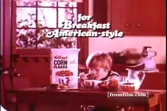 The still features a young boy sitting at a wooden table, enjoying a bowl of Kellogg's Corn Flakes. The background showcases a cozy kitchen setting with a window and cupboards. Prominent text above reads 'for Breakfast American-style,' emphasizing the product's appeal. The colors are warm and nostalgic, typical of 1970s advertising.