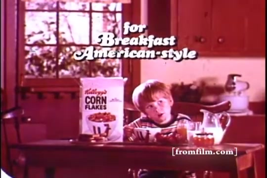 The still features a young boy sitting at a wooden table, enjoying a breakfast of Kellogg's Corn Flakes. A box of Corn Flakes is prominently displayed, alongside a glass of milk and a bowl. The scene is warm and inviting, with sunlight streaming through a window, creating a nostalgic, 1970s American breakfast atmosphere. The text 'for Breakfast American-style' is featured above the boy, emphasizing the advertisement's appeal.