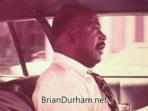 The still features a man seated in a car, likely captured during a moment of conversation or thought. He is dressed in a formal shirt and tie, reflecting a professional appearance. The background suggests urban scenery typical of Ohio in the 1970s, with a focus on the man's expression and demeanor. The color palette hints at the period's film quality, characterized by a soft, slightly faded look.