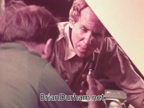 The still from 'Job Service PSA - Ohio - 1976' depicts two men engaged in a discussion in a workshop setting. One man is examining a small component, possibly for a vehicle, while the other attentively observes. The scene conveys a sense of hands-on skills and collaboration, reflecting the theme of job services in the context of mechanical work. The color tone suggests a vintage aesthetic typical of the 1970s.