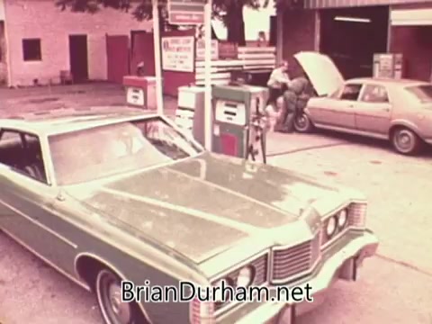 The still features a vintage gas station scene from 1976, showcasing a green car parked at a gas pump. A person is seen filling up the tank, while another individual leans against a car nearby, possibly engaging in conversation. The setting evokes a retro ambiance, characteristic of small-town America during that era, reflecting the theme of job services and community engagement.