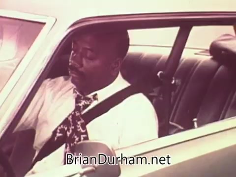 The still features a man seated in a car, wearing a white shirt and a patterned tie. He appears to be engaging with the vehicle's interior, likely in a contemplative state. The image is part of a public service announcement from Ohio in 1976, promoting job services. The overall tone reflects the era's aesthetics and themes related to employment and community support.