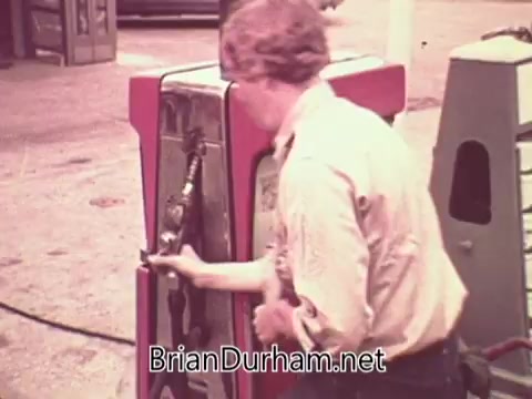 The still captures a young man at an old-fashioned gas pump in Ohio, circa 1976. He is dressed casually and is seen in the act of filling a vehicle with gasoline. The scene reflects the everyday life and work environment of the era, emphasizing job service and the automotive culture of the time. The colors and aesthetic are characteristic of 1970s film.