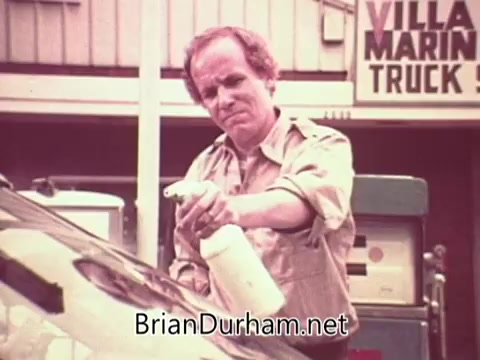 The still from 'Job Service PSA - Ohio - 1976' features a man spraying a solution onto a car windshield. He appears focused and determined, dressed in a short-sleeved shirt. In the background, there are signs indicating a truck service, suggesting the setting is a service station. The overall tone reflects a practical, hands-on approach to work.