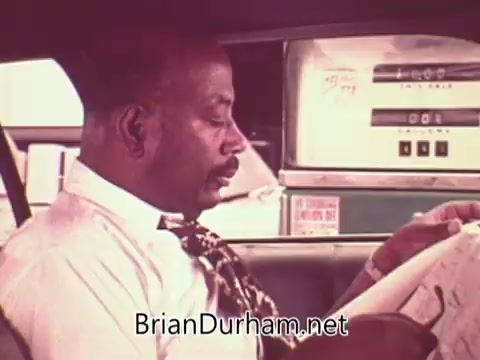 The still features a man dressed in a white shirt and patterned tie, sitting in a car and reading a newspaper. The setting suggests a moment of reflection or contemplation, likely related to employment or job searching, in the context of a public service announcement aimed at encouraging job-seeking efforts. The visual emphasizes a mundane yet significant aspect of navigating the job market in the 1970s.