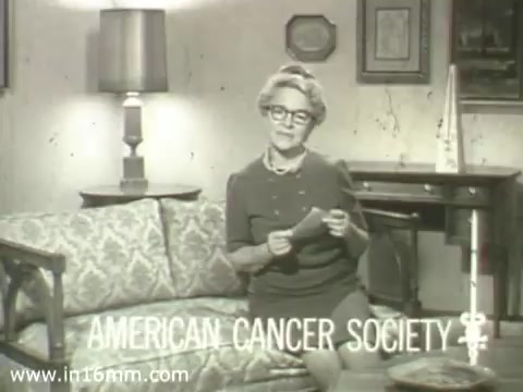 The still features a woman seated on a patterned couch in a home setting, delivering a public service announcement for the American Cancer Society from the 1960s. She appears to be middle-aged, wearing glasses and a modest dress, holding a piece of paper. The backdrop includes a lamp and framed artwork, evoking a domestic atmosphere. The text 'AMERICAN CANCER SOCIETY' is prominently displayed at the bottom, emphasizing the message of the PSA. The overall tone is earnest, aligning with the intent to raise awareness about cancer.