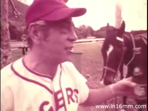 The still depicts Robert Strauss in a casual baseball-themed outfit, wearing a cap and a shirt that says 'GER.' He is animatedly speaking while holding a bottle of Canada Dry, seemingly in a relaxed outdoor setting with a horse in the background. The image has a warm, vintage tone typical of 16mm film.
