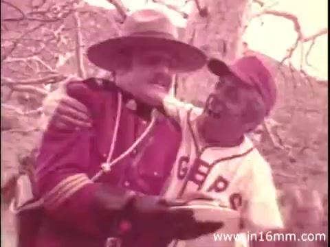 The still features two characters: one dressed as a Royal Canadian Mounted Police officer, wearing a wide-brimmed hat, and the other in a baseball uniform. They appear to be enjoying a jovial moment together, likely as part of a comedic exchange. The setting hints at an outdoor location with trees in the background, contributing to a lighthearted and friendly atmosphere typical of commercials. The color palette is muted, characteristic of older 16mm films.
