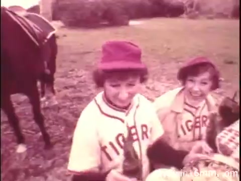The still from the Canada Dry commercial shows two young girls wearing baseball jerseys labeled 'TIGER,' enjoying a picnic. They are smiling and holding bottles, with a horse visible in the background. The muted color palette suggests it is an older print, typical of 16mm film. The setting appears casual and playful, emphasizing a lighthearted, nostalgic vibe associated with the product.