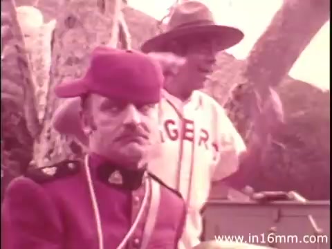 The still from the Canada Dry commercial features two characters: one is an austere-looking man in a red military uniform, while the other, dressed casually in a baseball jersey and hat, appears animated and engaging. The background suggests a natural setting, contributing to the lighthearted tone typical of commercials from that era. The visual quality indicates it's from a vintage 16mm print.