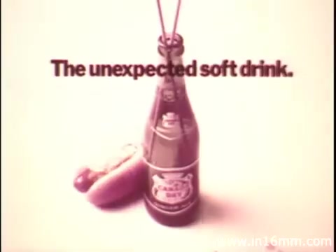 The still features a bottle of Canada Dry soda prominently displayed, alongside a hot dog, conveying an unexpected pairing. The text 'The unexpected soft drink.' overlays the image, suggesting a surprising choice for a beverage. The overall aesthetics reflect a vintage advertising style typical of that era.