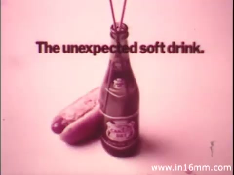 The still features a bottle of Canada Dry positioned prominently in the foreground, alongside a hot dog or sausage. The text 'The unexpected soft drink.' is displayed above the items. The overall color scheme is soft and muted, likely giving a vintage feel characteristic of 16mm film.