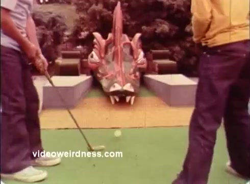 The still features two golfers preparing to take a shot at a miniature golf course adorned with a large, colorful dragon-like sculpture. The vibrant setting likely adds a whimsical touch to the commercial, which connects to the Dinah Shore Golf Tournament, emphasizing a playful, family-friendly atmosphere.