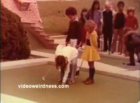 The still from the 1970s Colgate TV commercial features a group of children on a miniature golf course. One child is crouching down, possibly preparing to putt, while another child stands nearby, observing. In the background, several children watch, creating a playful scene reminiscent of youthful competition. The setting appears sunny and casual, aligning with the lighthearted spirit of the Dinah Shore Golf Tournament promotions.