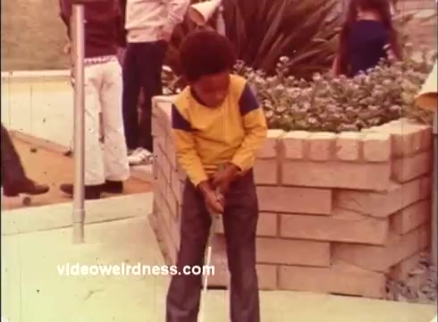 The still features a young boy dressed in a yellow and blue long-sleeve shirt, poised with a golf club, seemingly preparing to make a shot. He's surrounded by a casual outdoor setting with a brick wall and foliage, hinting at a festive atmosphere. In the background, other people are engaged, possibly emphasizing the context of the Dinah Shore Golf Tournament from the 1970s. The commercial likely promotes Colgate in a playful, family-friendly manner.
