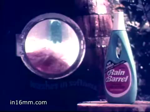 The still features a vintage bottle of Rain Barrel Fabric Softener prominently positioned next to a rustic wooden rain barrel. A circular window on the barrel reveals soft, billowing fabric inside, reinforcing the theme of softness. The color palette is vibrant, typical of 1970s advertisements, with the bottle's branding clearly visible. The scene conveys a nostalgic and natural approach to fabric care, emphasizing the product's beneficial qualities.