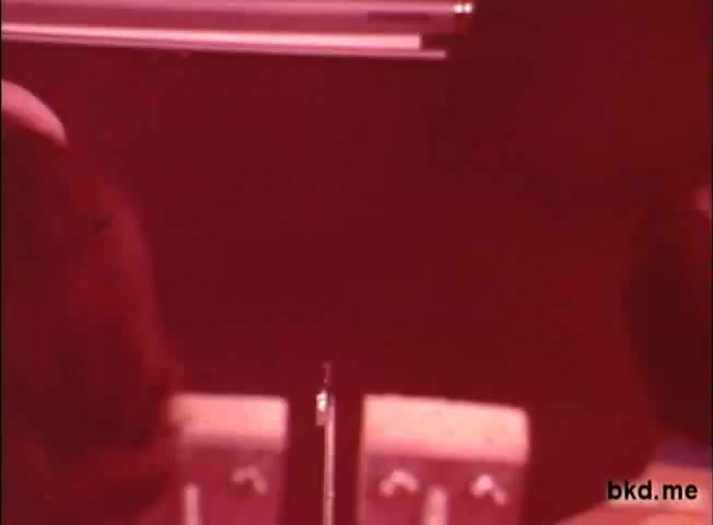 The still from the 'Spic and Span' TV commercial features a dimly lit scene, predominantly bathed in a red hue. It captures two individuals, partially visible, possibly engaging in conversation. The background includes indistinct objects, likely appliances or cleaning products, with a low level of detail typical of vintage 16mm film. The overall atmosphere conveys a nostalgic and retro aesthetic reminiscent of advertisements from the 1960s to 1970s.