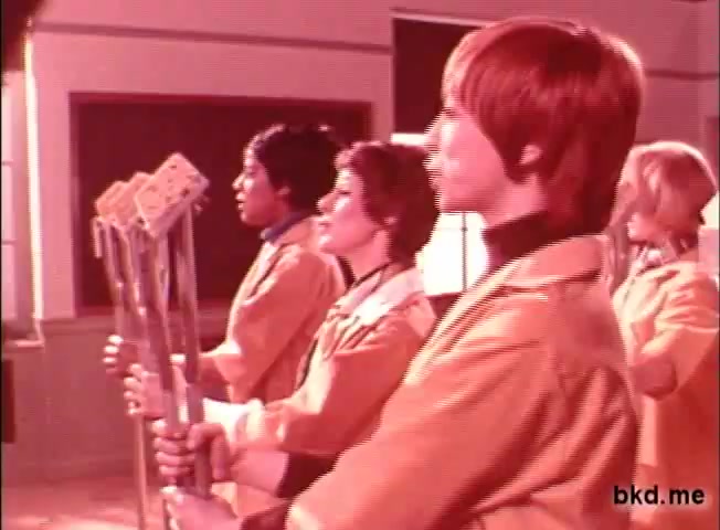 The still features four individuals standing in a line, each holding cleaning tools, likely mops. They are dressed in vibrant yellow or orange coats, suggesting a playful or energetic theme typical of 1960s-1970s advertisements. The background is simple, emphasizing the characters and their tools, likely promoting the Spic and Span cleaning product in a lighthearted manner. The overall tone conveys a fun approach to cleaning.