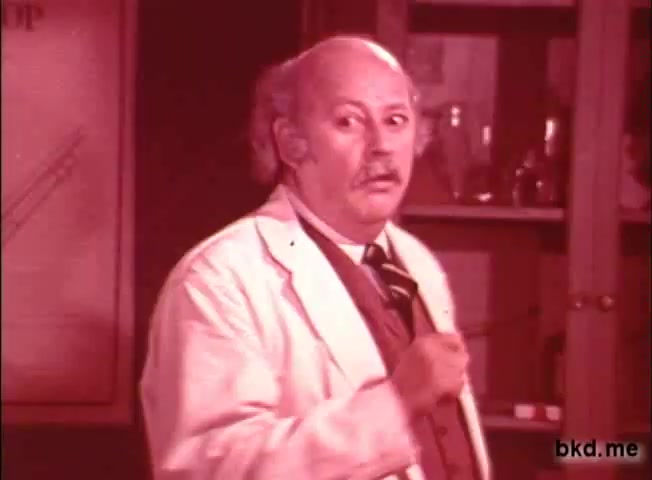 The still features a man in a white lab coat, likely portraying a scientist or expert, with a concerned expression. He gestures with a pointer in hand, suggesting he is explaining something important. The background consists of shelves with glassware and laboratory equipment, enhancing the scientific context. The color tone is slightly muted, typical of the 1960s-1970s film style.
