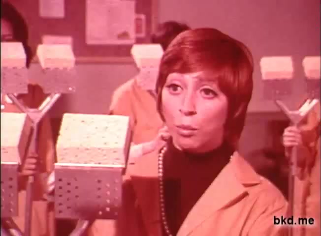 The still features a woman with short hair, dressed in a mustard-colored coat and a black top, addressing the camera while holding a kitchen sponge. She appears to be in a classroom or training environment, with individuals in similar attire positioned behind her, all holding sponges. The color palette is characteristic of the late 1960s to 1970s era, suggesting a promotional context for the Spic and Span brand. The overall tone is instructional and lively, typical of television commercials from that period.