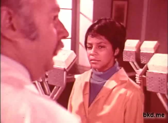 The still from the 'Spic and Span' TV commercial features two characters: a man with a mustache and a woman wearing a yellow lab coat. The woman appears to be listening intently to the man, who is talking. The background suggests a bright, professional setting, likely a laboratory or cleaning environment, with equipment visible behind them. The color palette has a muted yet warm tone typical of 1960s-1970s commercials.