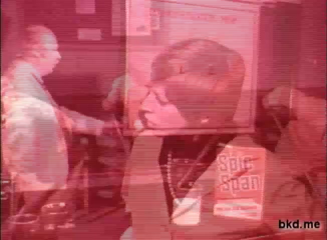 The still from the 'Spic and Span' TV commercial features a vibrant, retro atmosphere typical of the 1960s-1970s. A woman with a short haircut is in focus, appearing engaged in conversation. The background contains figures and kitchen-related elements, hinting at a domestic setting. The color palette has a red tint, likely due to the vintage film print, giving it a nostalgic feel. The commercial seems to emphasize cleanliness and home life, central themes of the brand.