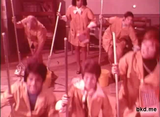 The still features a group of children energetically engaged in a cleaning activity, dressed in bright yellow raincoats. They wield brooms and mops, creating a playful and chaotic atmosphere. The background suggests a classroom setting, while the overall color tone has a warm, reddish hue, typical of 1960s-70s film prints. The scene conveys a sense of fun and teamwork, likely promoting the Spic and Span cleaning product.