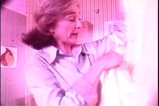 The still from the 1970s Downy Fabric Softener commercial shows a woman with light brown hair, expressing excitement as she holds up a fabric or garment. The scene is brightly lit, with pastel colors in the background, giving it a vintage aesthetic typical of the time. The emphasis is on the softness and freshness of the fabric, which is central to the product's appeal.