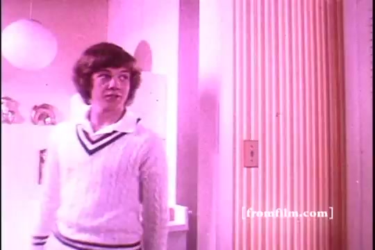 The still features a young male character dressed in a white sweater with a navy stripe, standing in a brightly lit, pastel-colored room typical of the 1970s aesthetic. He appears to be engaged in a moment of curiosity or surprise, likely in response to the presence of Downy Fabric Softener in the context of the commercial. The background includes retro decor, enhancing the nostalgic feel of the era.