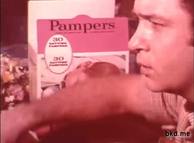 The still shows a man closely interacting with a pack of Pampers diapers prominently displayed in the background. The packaging features a bright, colorful design typical of the 1970s, highlighting the quantity of diapers. The man's expression appears engaged, possibly demonstrating or discussing the product. The overall tone reflects the nostalgic advertising style of the era.