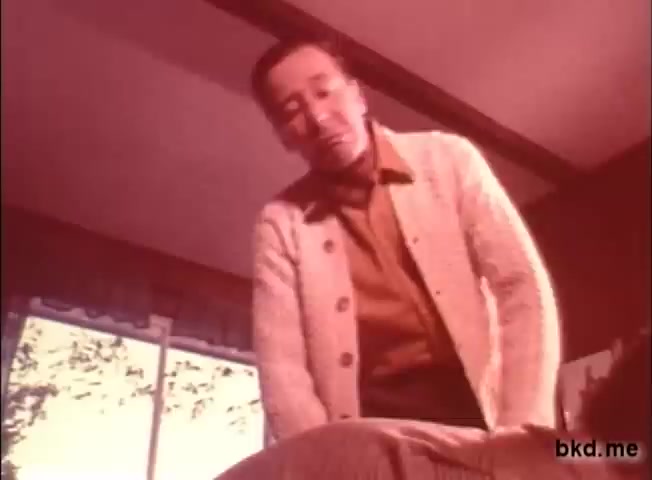 The still features a middle-aged man wearing a light-colored cardigan over a brown shirt, leaning over to interact with an off-screen subject, likely a child. The setting appears warm and homey, with soft lighting filtering through a window. The nostalgic color tone of the image reflects the 1970s aesthetic, capturing the essence of a family-oriented theme typical of diaper commercials from that era.