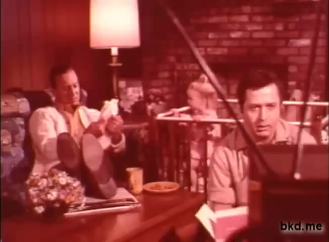 The still features a cozy 1970s living room setting, with two men sitting in front of a television. One man is relaxed with his feet up, holding a diaper, while the other appears focused on the TV. A child is visible in a crib nearby, and there are domestic elements like a lamp and snacks on the table. The color tone is warm, typical of the era's film quality.