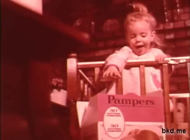 The still features a young girl joyfully leaning over a crib railing, with a box of Pampers diapers prominently displayed in the foreground. The color tone is warm and nostalgic, typical of 1970s commercials. The setting appears cozy, emphasizing family and childhood themes associated with the product.
