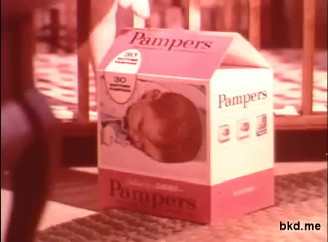 The still shows a 1970s Pampers diaper box prominently featuring a colorful design typical of that era. The box has a large image of a baby's head on the front and is labeled with the brand name 'Pampers.' The scene seems to be set in a nursery, with wooden bars of a crib in the background, suggesting a focus on baby care products. The overall tone is warm and nostalgic.
