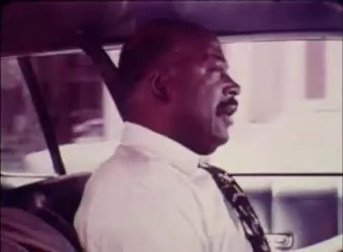 The still from the 16mm print of the public service commercial for Job Service features a man in profile, seated in a car. He has a mustache and wears a white shirt with a tie, conveying a professional appearance. The interior of the car is visible, suggesting a focus on communication or travel related to job placement. The overall tone of the commercial is informative, promoting the use of Job Service to assist in filling job openings for the Colorado Division of Employment.