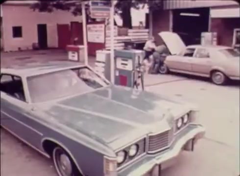 The still features a vintage scene typical of the late 1970s to early 1980s, showcasing a green car parked at a gasoline station. In the background, a man is filling up a tank while another vehicle is visible with its hood open, suggesting maintenance or repairs. The setting has a nostalgic feel, with classic gas pumps and a modest service station, reflecting the era's roadside culture. This imagery aligns with the commercial's aim to promote Job Service as a valuable resource for filling job openings in the community.