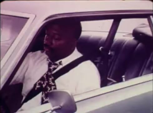 The still from the 16mm print shows a man seated in a car, wearing a white shirt and patterned tie, with a seatbelt fastened. The setting suggests a focus on employment and job services, relevant to the public service commercial for Job Service by the Colorado Division of Employment. The overall tone conveys professionalism and readiness to engage in job-seeking activities.