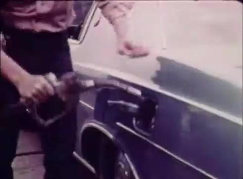 The still from the 'Job Service' commercial shows a person filling a car with gasoline at a fuel pump. The scene captures a moment of everyday life, emphasizing the message of the commercial about job services assisting with employment needs. The visual style reflects the vintage aesthetic of 16mm film, typical of the period when the commercial was made.