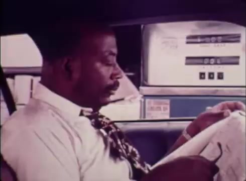 The still from the public service commercial for Job Service features a man seated in a car, focused on reading a document, possibly a job listing. The environment suggests he is at a gas station, with pumps visible in the background. His attire is formal, which indicates a professional context. The overall tone aligns with the campaign's goal of promoting job placement assistance.