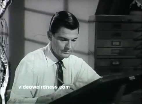 The still from 'Kaiser Aluminum - The Bright Idea' features a man in a white dress shirt and tie, focused on drawing or drafting at a desk. The background includes a hint of horizontal lines, possibly from a window. The scene conveys a sense of professionalism and innovation, aligning with the theme of using aluminum for better living at lower costs. The print appears to have some wear, typical of vintage 16mm film.