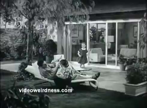The still from 'Kaiser Aluminum - The Bright Idea' depicts a serene domestic scene in a backyard. Two men are lounging on a deck chair, relaxed and engaged in conversation, while a young girl plays nearby. The setting features a modern home with large glass doors opening to the exterior. This image reflects the theme of better living and the cost-effective advantages of aluminum, emphasizing a comfortable lifestyle facilitated by modern materials.