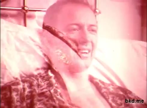 The still from the 'Mentholatum commercial - 1970's' features a man in a leopard print robe, smiling while reclining on a bed. The image has a soft, slightly faded quality typical of 16mm prints, with a warm color tone emphasizing a nostalgic feeling. The composition suggests a relaxed, casual atmosphere, likely aimed at conveying comfort and satisfaction associated with the product.
