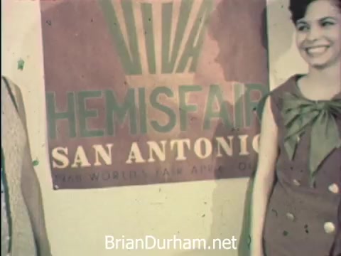 The still captures a moment from 'News Item - Secretaries at Hemisfair '68,' showcasing two women smiling in front of a colorful banner promoting Hemisfair in San Antonio. It reflects the celebration of Secretaries' Week, highlighting the blend of work and leisure as they navigate their responsibilities amidst the vibrant atmosphere of the world's fair, with the backdrop hinting at the festivities and activities available to them.